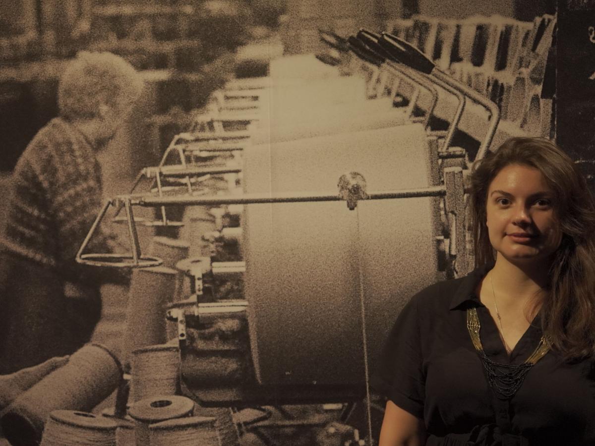 A photo of Carla Sayer in front of a wall display showing an archive image of two people working machinery.
