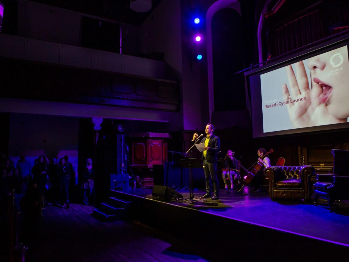 A photo of Gareth on stage at the launch event, The Breath Cycle name is on a big screen behind them.