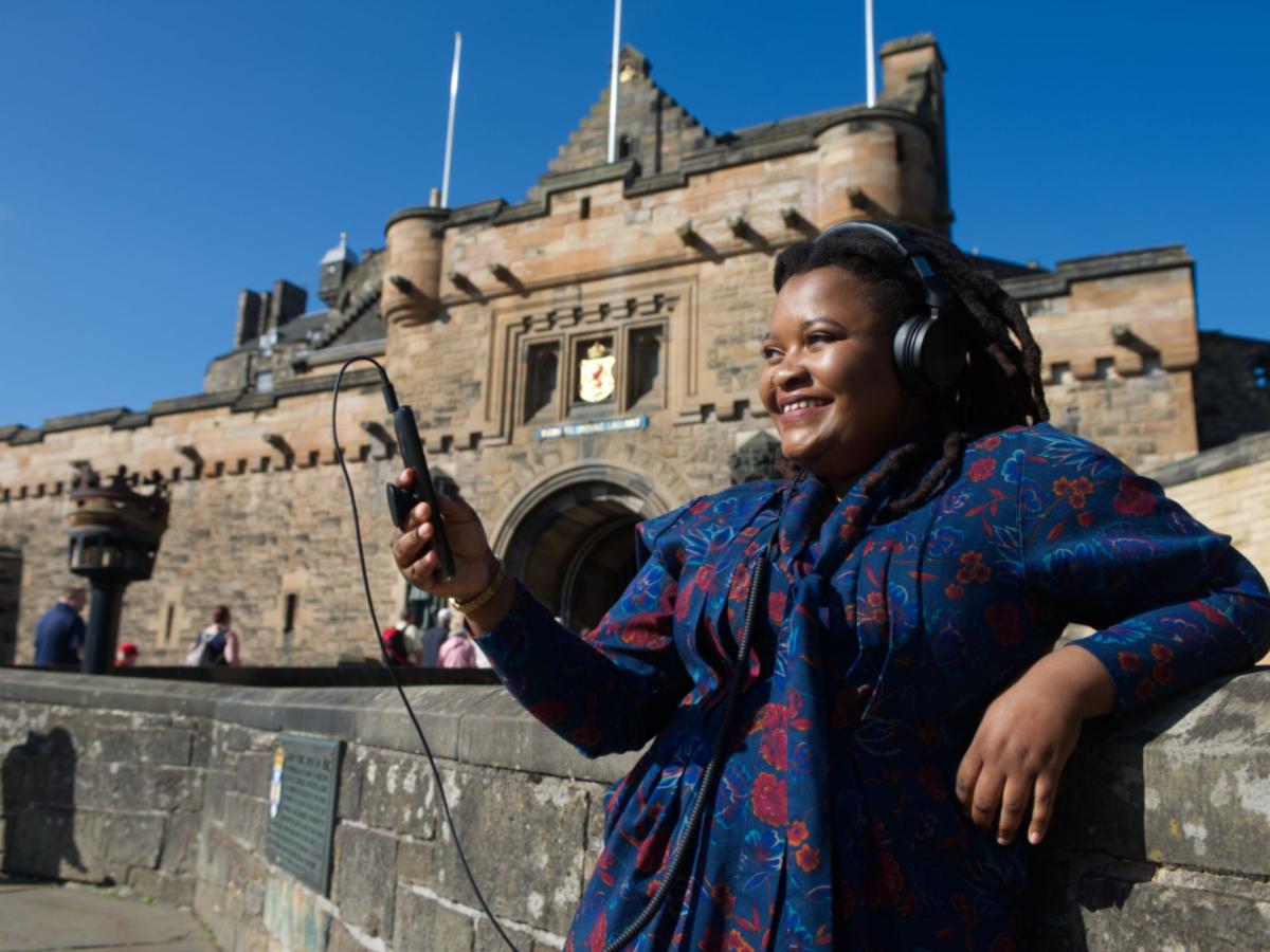 A photo of Tanatsei Gambura with a headset on in front of Edinburgh Castle.