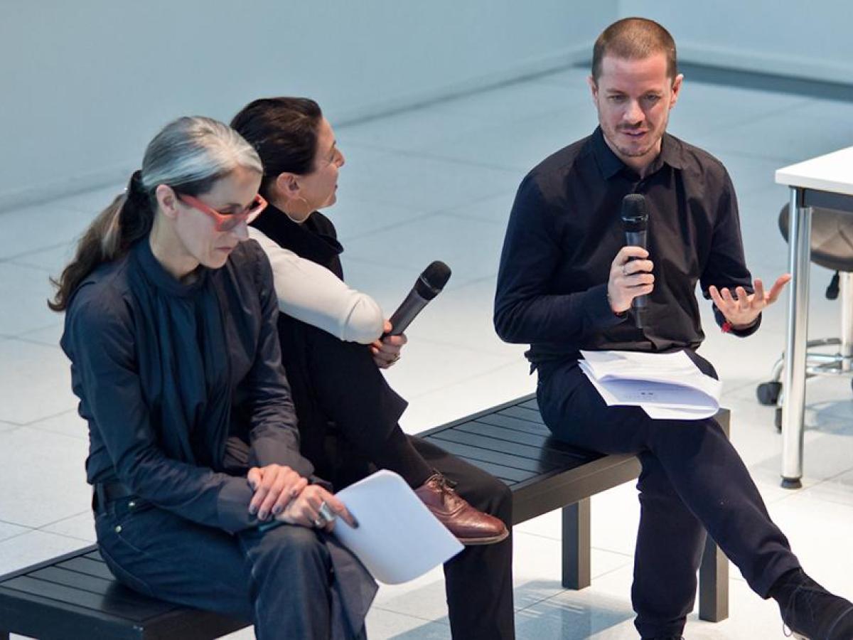 Three people sat on a bench, chairing an event. Two are holding microphones, with one person speaking into one.