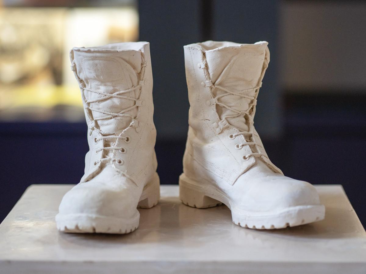 A cast of a pair of combat boots. They're unlaced and slightly worn; an off-white colour standing on a marble pedestal