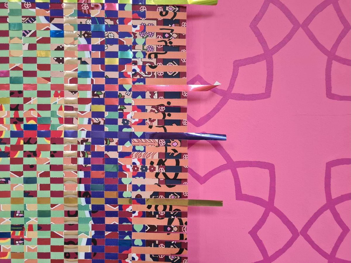 The left side of the artwork is made up of woven paper strips of various colours. The right side has a dark pink pattern on a pale pink background