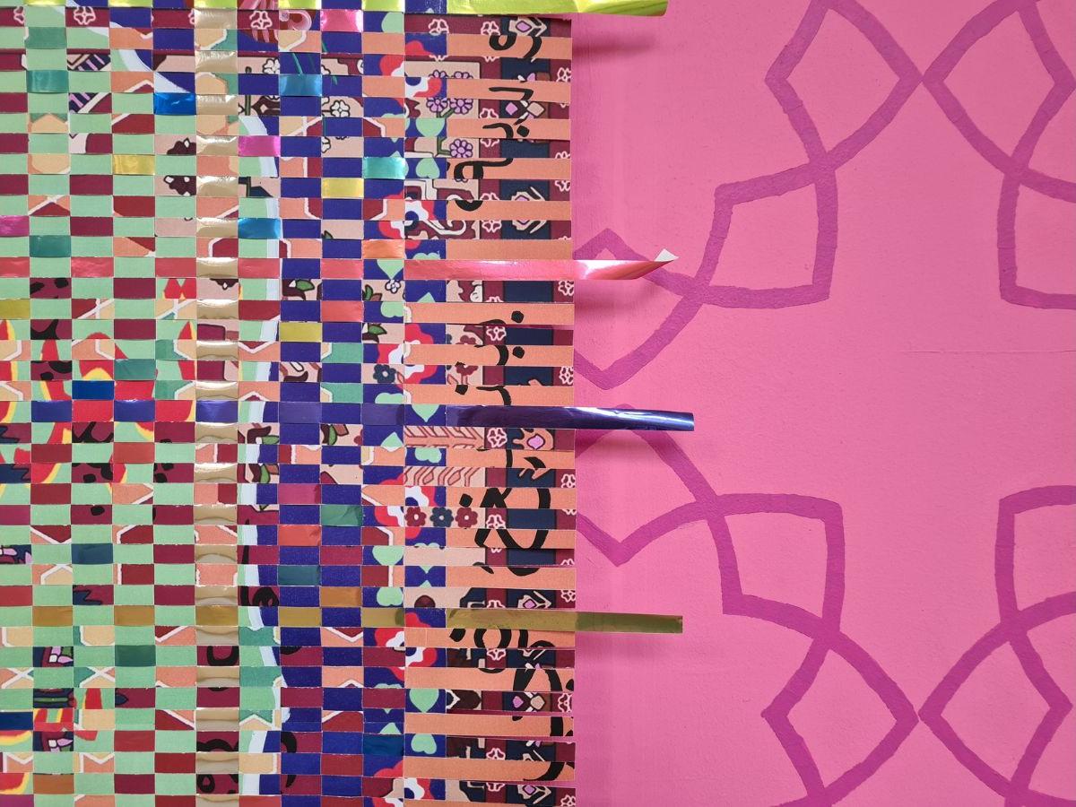 The left side of the artwork is made up of woven paper strips of various colours. The right side has a dark pink pattern on a pale pink background