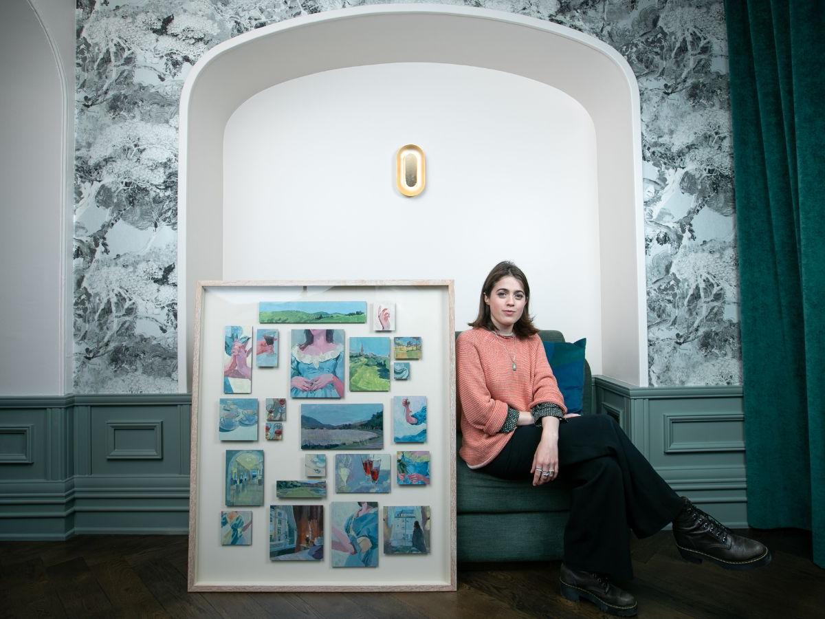 Ella Williams sits beside her framed artwork. The work consists of numerous small paintings, in a large white frame. 