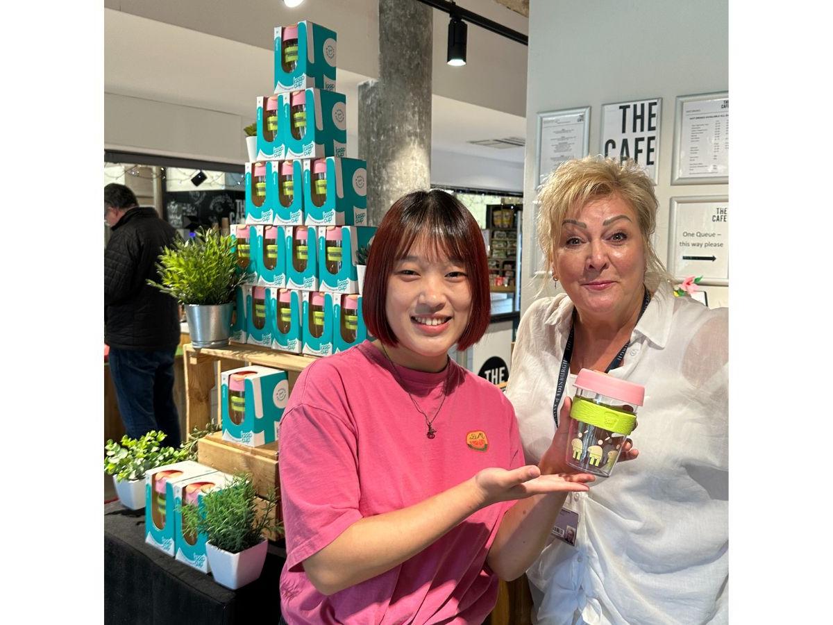 Ruge is smiling, holding up her KeepCup, which has a pale pink lid and a pale green band around it. Lita stands next to her. Behind them is a pyramid of KeepCups in their boxes.