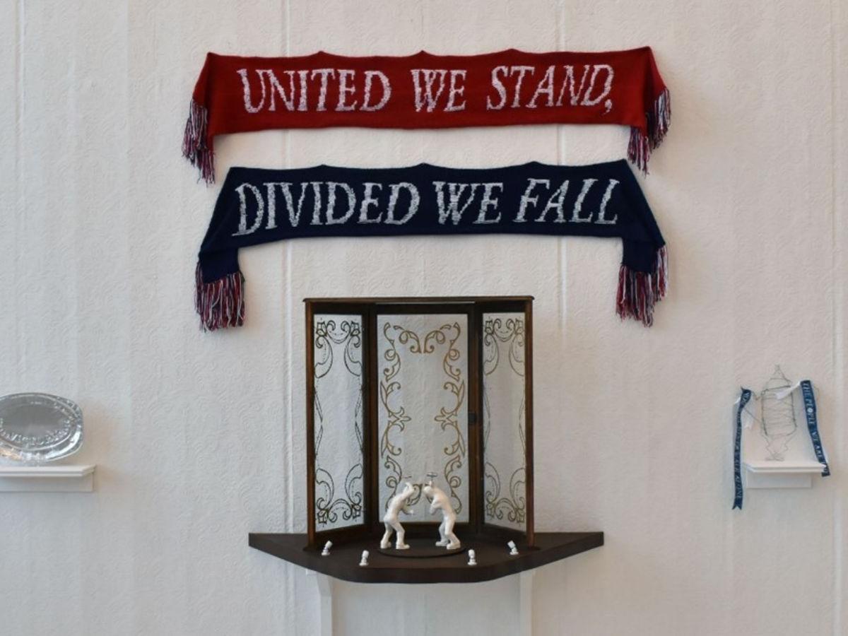 A white gallery wall. At the top are pinned two scarves with white text on. The top one is red and reads 'United we stand,' the bottom one is blue and reads 'Divided we fall'. Beneath is a shelf holding a 3D frame and two white figures. There are smaller shelves either side, one holding a silver-coloured platter, the other a trophy with blue ribbons from the handles.