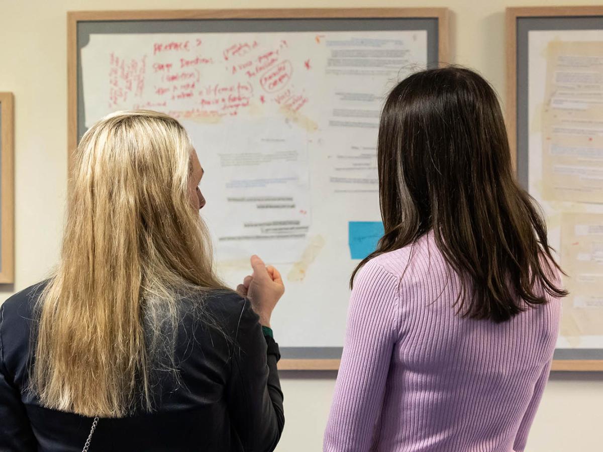 Two people with long hair have their backs to us. They are looking at a collage which is in a large wooden frame and hung on a wall.