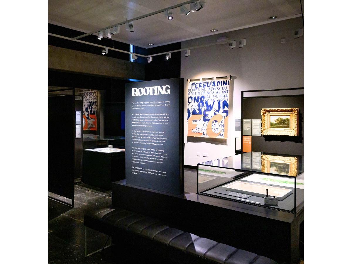 Interior shot of the Rooting exhibition - a black panel bears white text, headed Rooting, there are paintings and display cases behind.