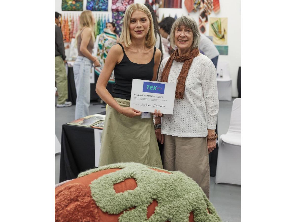 Winnie and Joanna stand together, Winnie holding a certificate. In front of them is a terracotta fabric shape, with textured green fabric patterns on