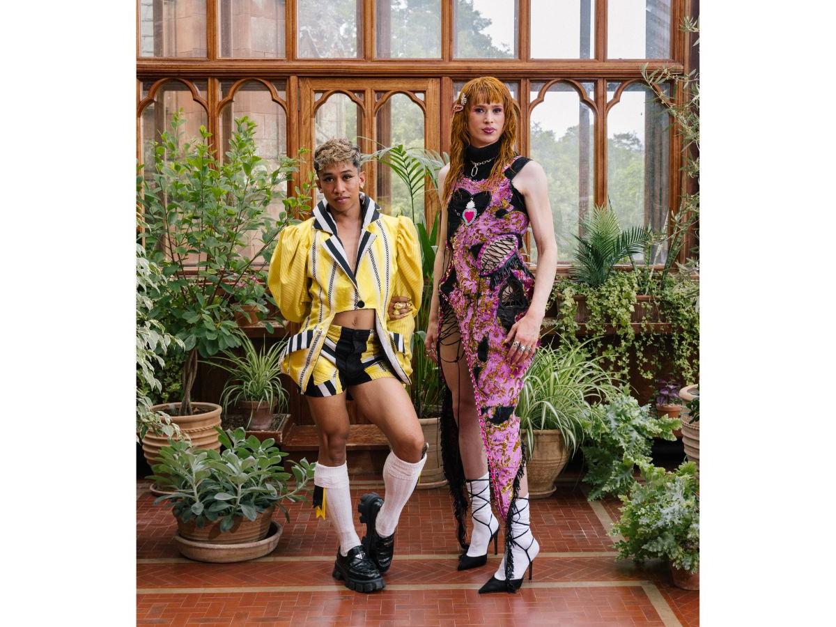 Davide and Angel stand in front of a wall of windows, lush green plants either side of them. Davide wears an elaborate yellow jacket and shorts, Angel a pink and black dress with black lacing.