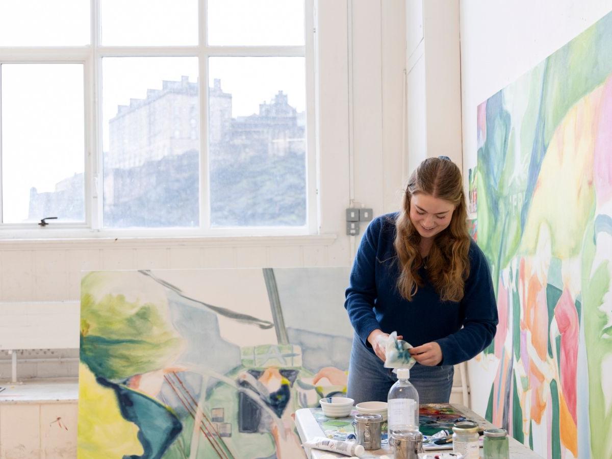 Phoebe in her studio at ECA. A colourful painting is on the wall beside her, another on the floor behind her. She looks to be cleaning brushes. The Castle is visible through the window behind her.