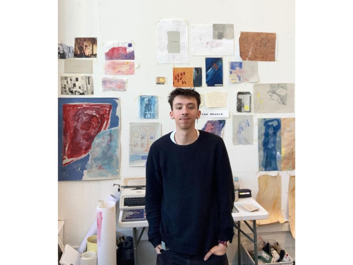 Thomas, wearing a black jumper, stands in his studio, looking at the camera. The white walls are adorned with paintings on sheets of various sizes. Behind him is a table, with a typewriter on it.