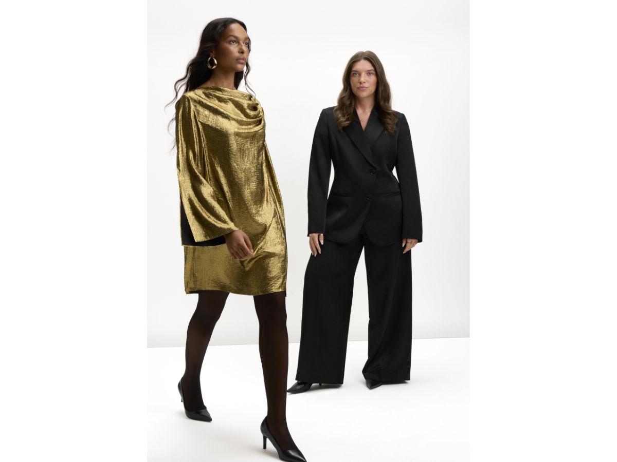 Against a white backdrop, Casey, wearing a black suit, stands as a model walks past, wearing her gold draped mini dress.