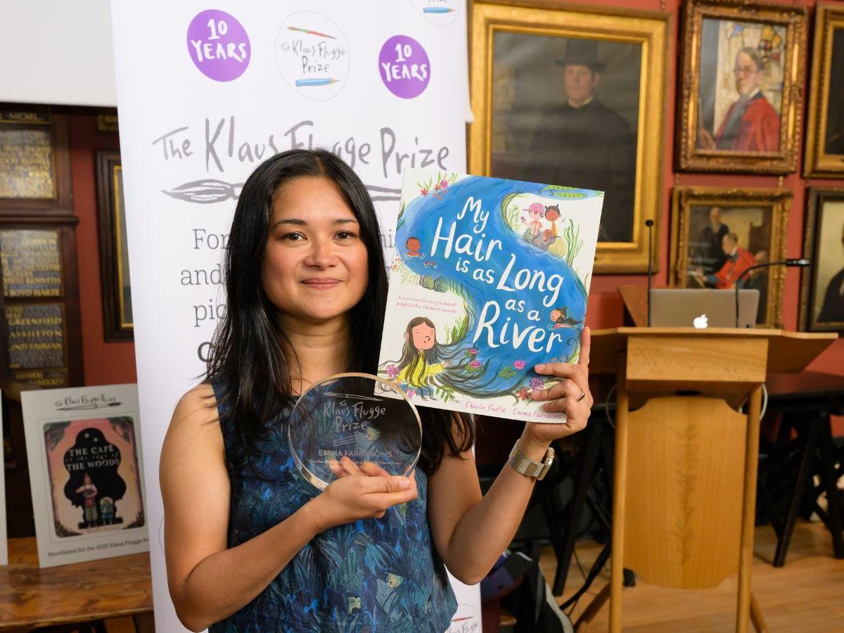 Emma holds a copy of the book, and her glass award. On the wall behind her are several framed portraits.
