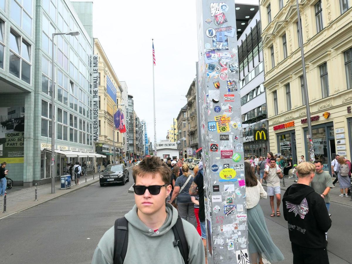 Matt, wearing sunglasses, a grey hoodie and a backpack, stands in the middle of a busy shopping street. He is next to a tall metal pole covered in stickers.