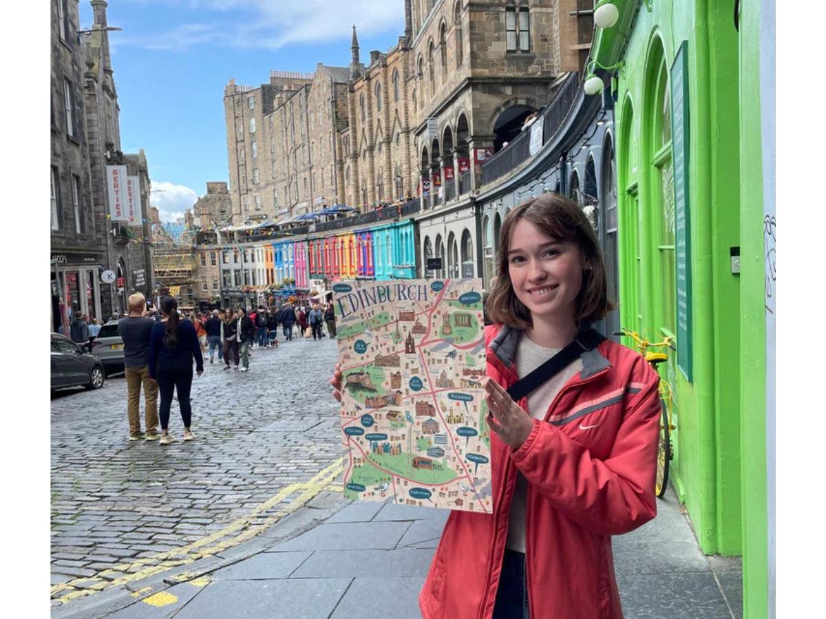 Ally stands in front of the coloured shop fronts of Victoria Street in Edinburgh, holding up one of her illustrated maps