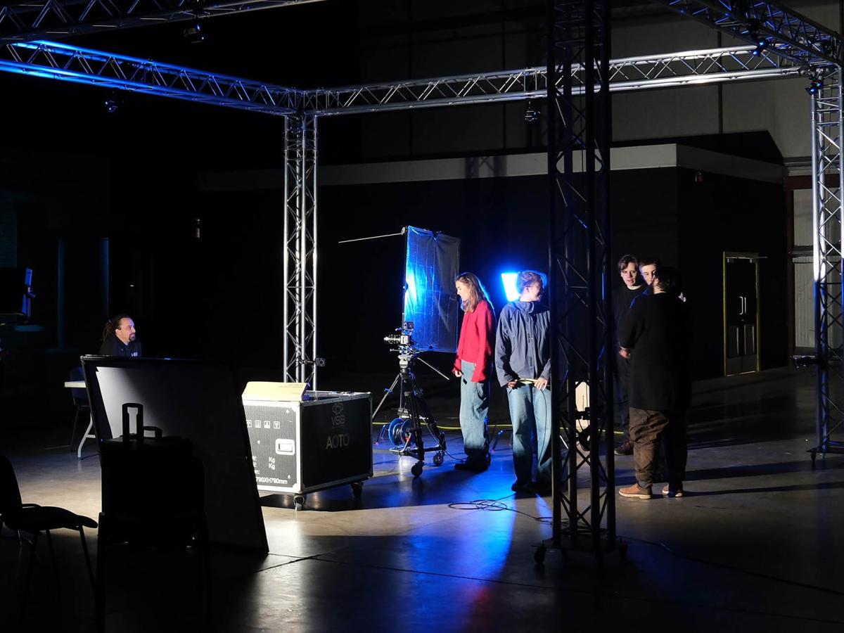 A small group of students are working with camera equipment. The studio is mainly dark but there are some dramatic spotlights.