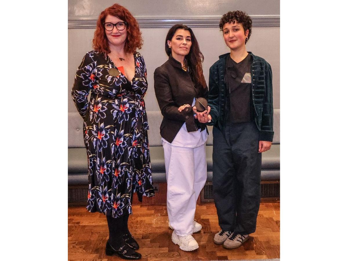 Tracy, Es and Rosina stand in a line. Es and Rosina cradle one award in their cupped hands - a metal circle with intersecting rectangle. Tracy is wearing her award - a circular brooch with a funky red magnetic backing visible at her neckline.