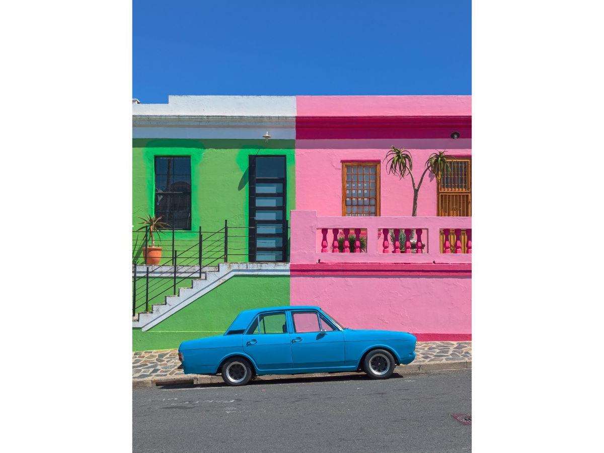 A photograph of a bright blue car parked in the street. Behind it are two buildings - the left is bright green, the right is bright pink. The dividing line between the two houses lines up exactly with the line between the car's front and rear side windows. A bright blue sky is above, the grey road below.