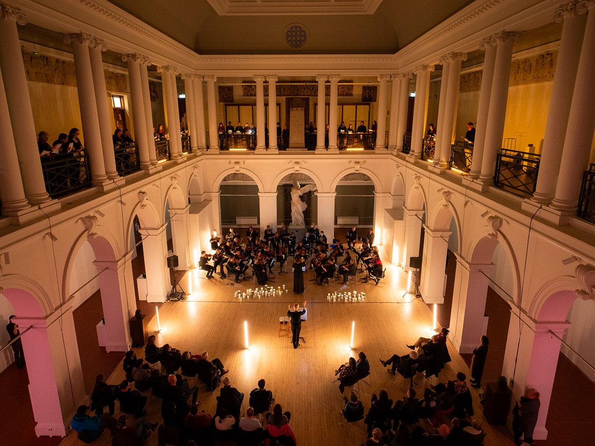 The Sculpture Court, beautifully lit. At one end is an orchestra; standing along the balcony on three sides of the room, is a choir. The conductor's arms are raised.