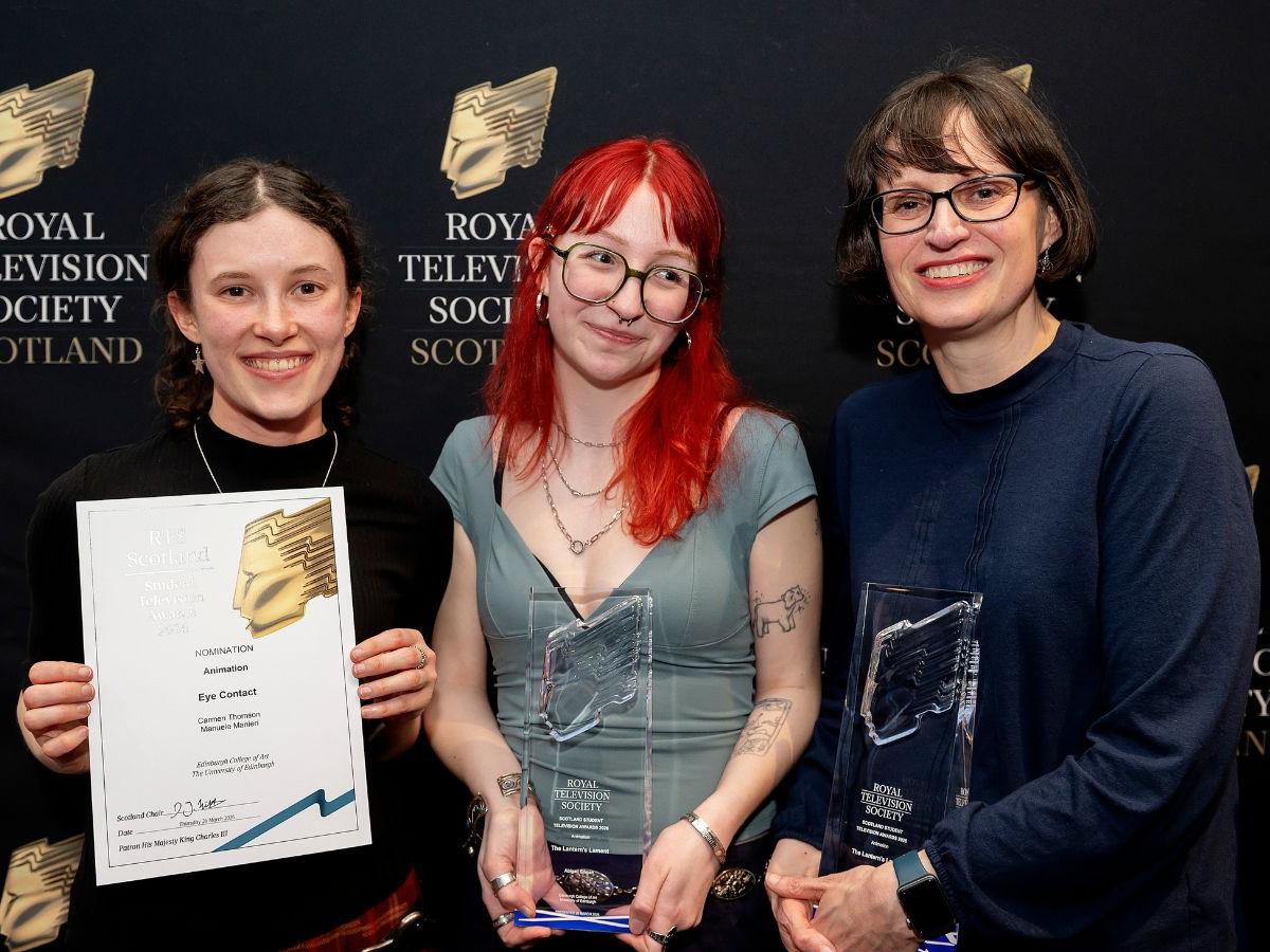 Carmen Thomson, Abby Gilman and Dr Nichola Dobson stand in front of a black board with a logo repeated across it – a gold shape like an angular, abstracted face in profile, and the words Royal Television Society Scotland.