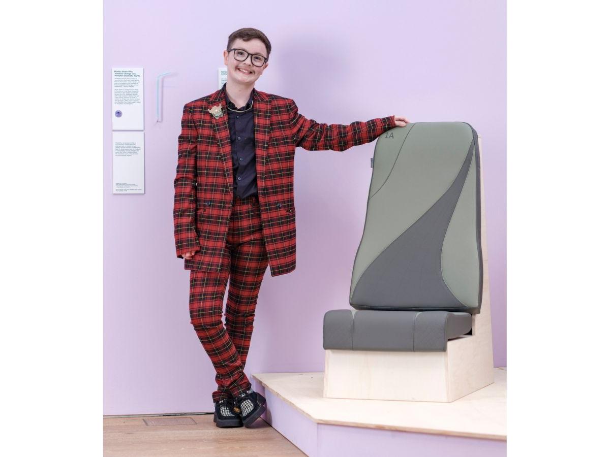 Jamie stands next to his airline seat. He is smiling, and is wearing a red and black tartan suit. The airline seat is a greenish grey, with a design including two curved lines, with a darker colour between them.