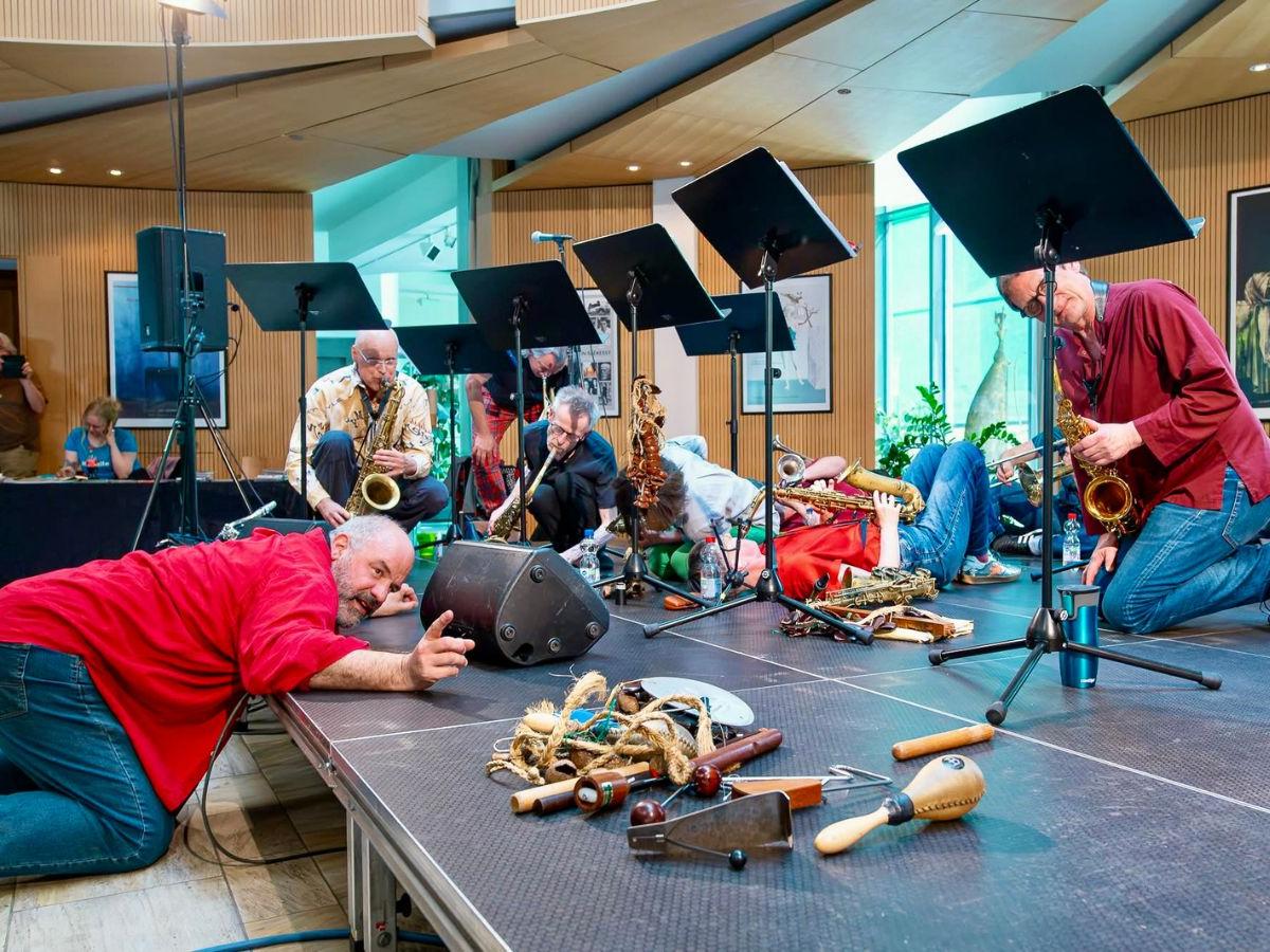 Raymond kneels on the floor in front of the stage, leaning onto the stage. On stage, five saxophonists are playing, all in different positions - kneeling, crouching, lying down. Also on stage are a collection of small instruments, and a row of music stands.