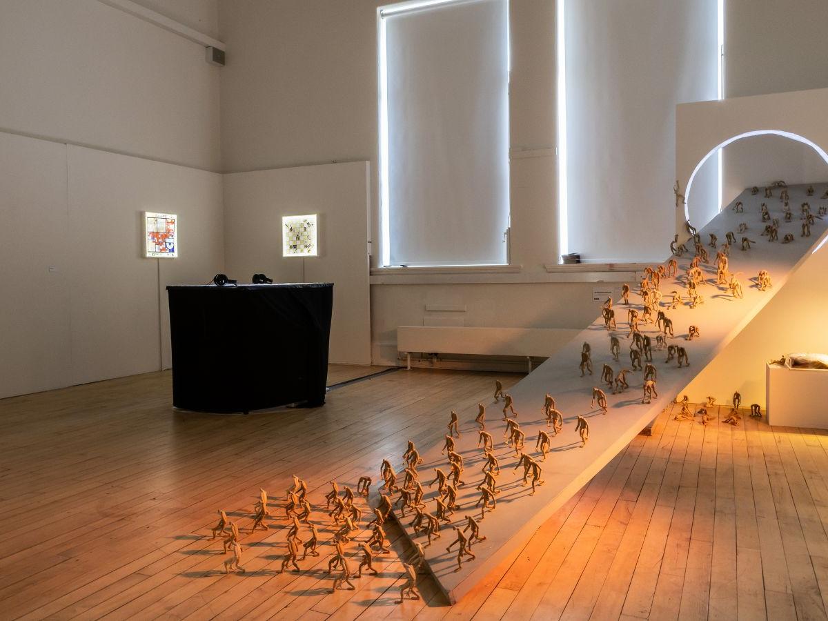 A studio at the Graduate Show, with low lighting. A ramp extends from the floor up to an illuminated circle. Lots of tiny figurines are descending down the ramp.