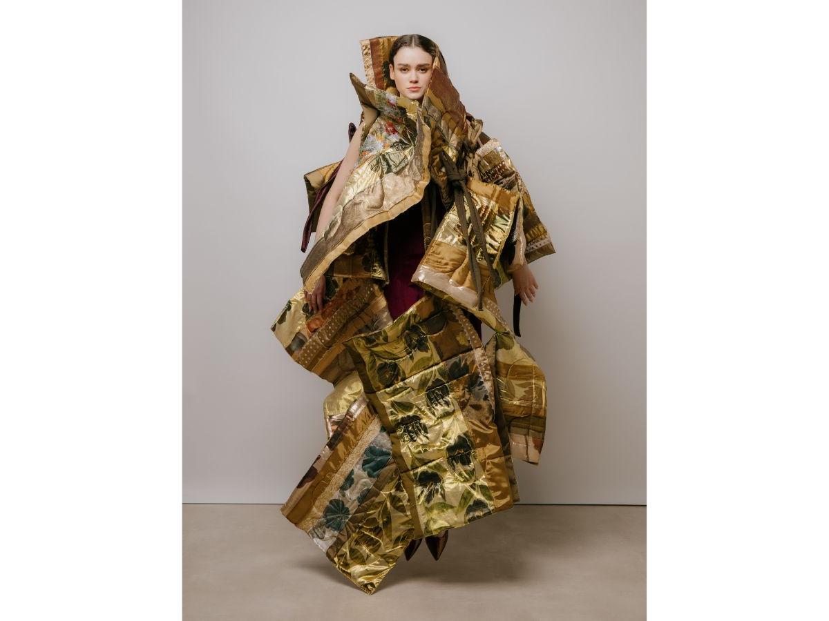 A model wears an outfit made of quilted golden patterned panels, hanging in a plethora of shapes and angles. She is jumping, both toes clear of the ground.