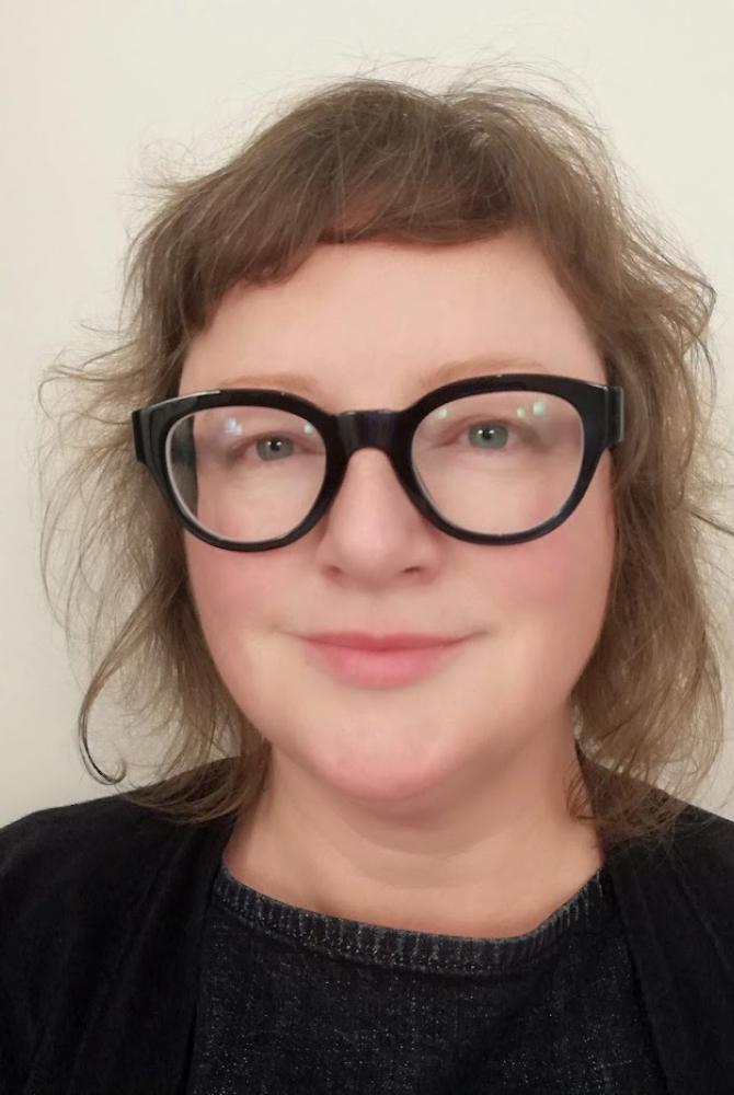 A person with short, light brown hair, wearing black rimmed glasses and smiling at the camera.