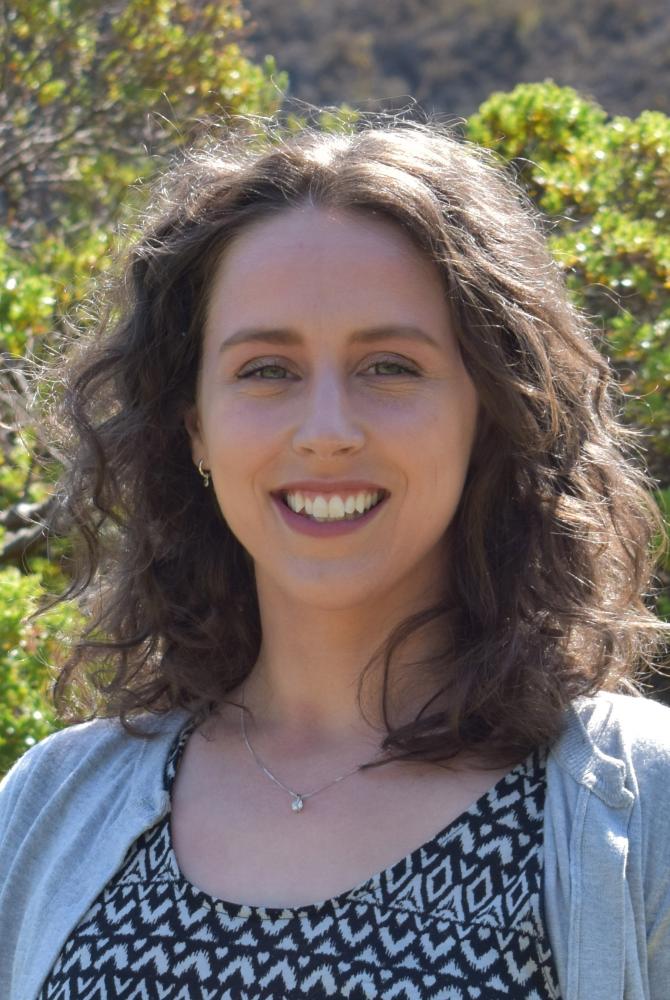 A person with shoulder length, brown curly hair, standing out in the sun and smiling