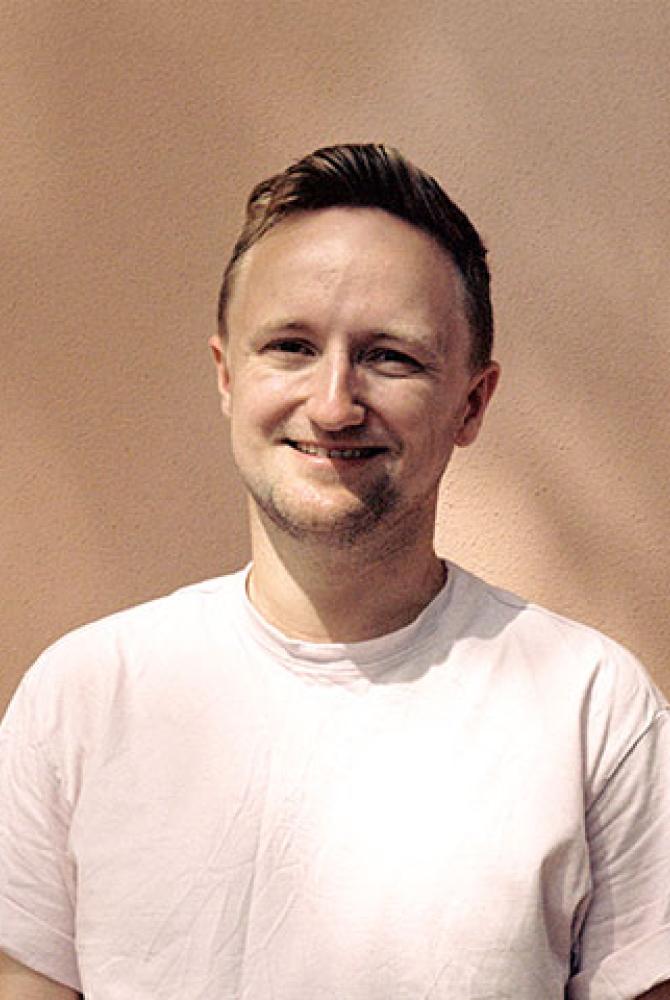 A person in a white t-shirt, standing against a light bisque coloured wall. The sunlight filters through the branches of a tree on to them.