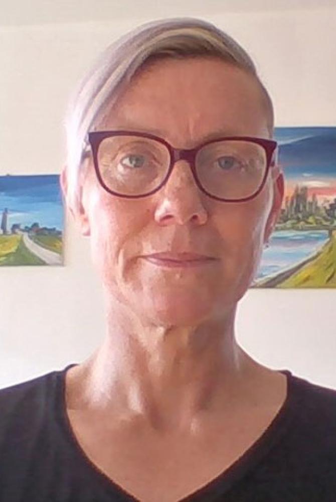 A person with short, silvery hair, wearing pink glasses and a dark top. They are looking directly to the camera, and have two paintings of landscapes hung up on the wall behind them.