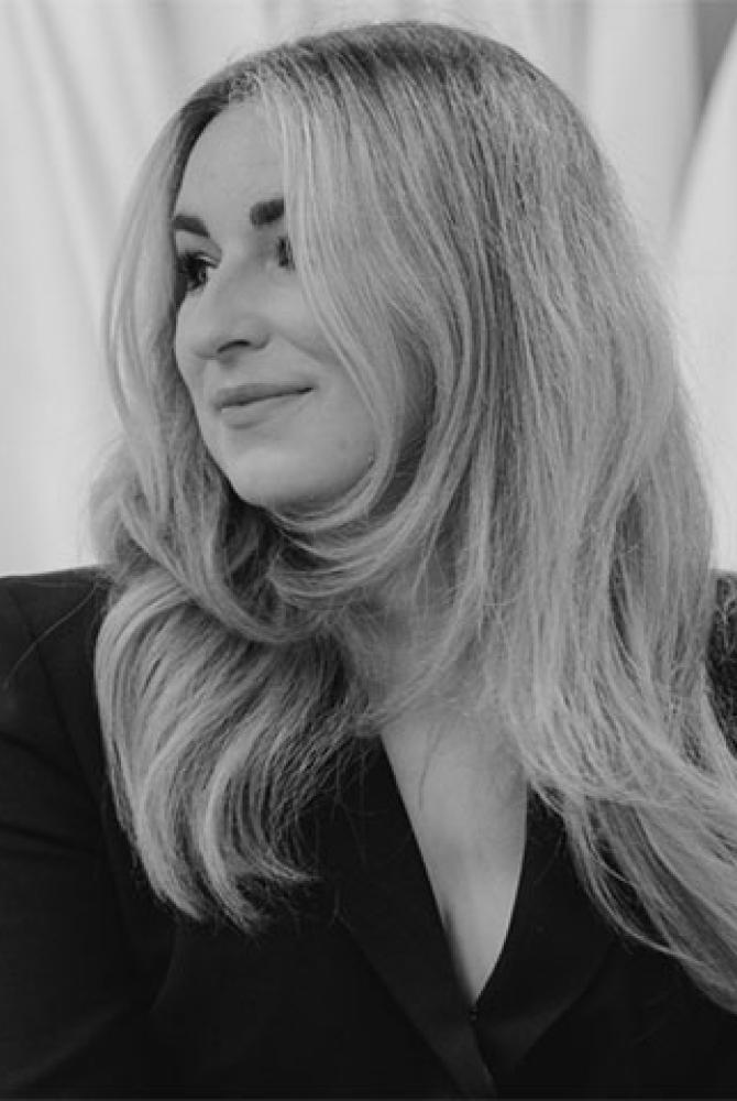 A black and white image of a person with long light coloured hair, looking sideways and smiling. They're wearing a dark coloured blazer.
