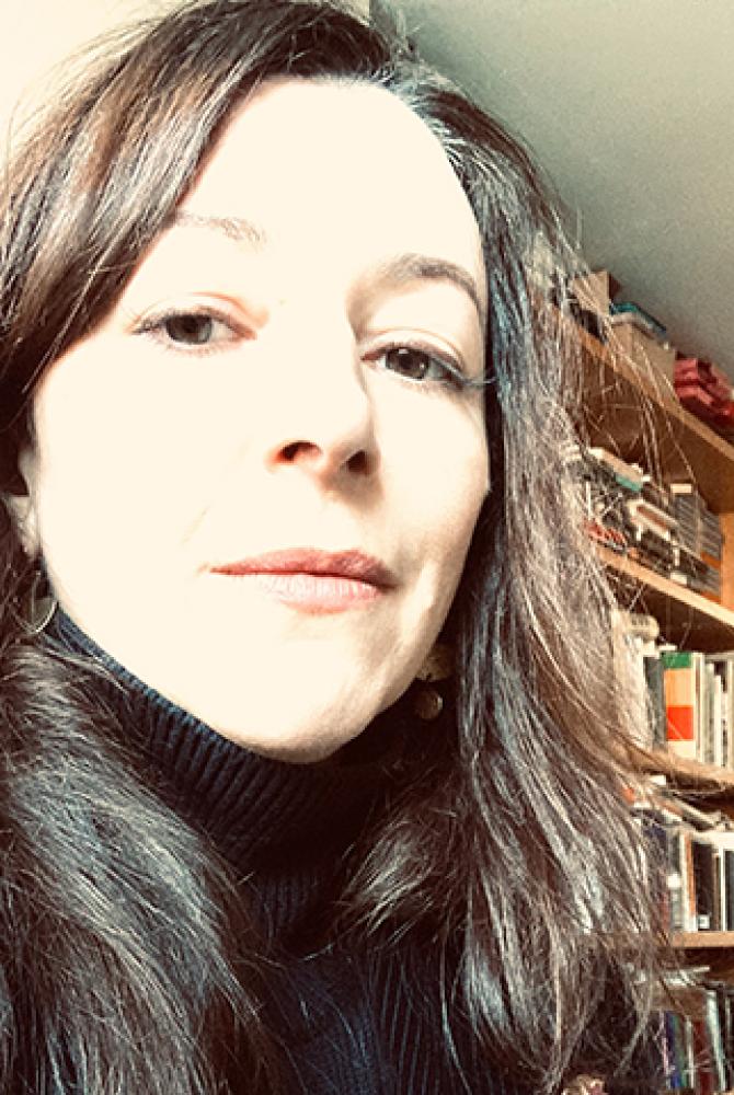 A person with long, dark hair, looks to the camera. There is a book case just behind them in the corner of the room.