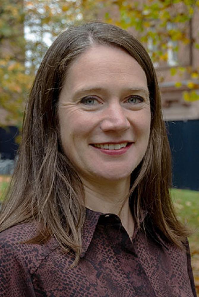 A person with long, light brown hair smiles to the camera. They're standing in the ECA courtyard, where the tree in the background is coming in to autumnal bloom.