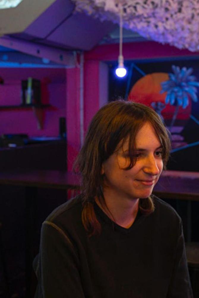A person with dark hair, sits in a room with red walls. There is a bare lightbulb in the background and a white, decorative hanging on the ceiling.