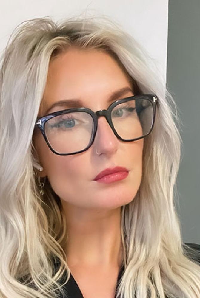 A person with long, blonde/silver hair, looking at the camera. They wear big, black glasses and are standing in front of a wall with a piece of white paper on it.
