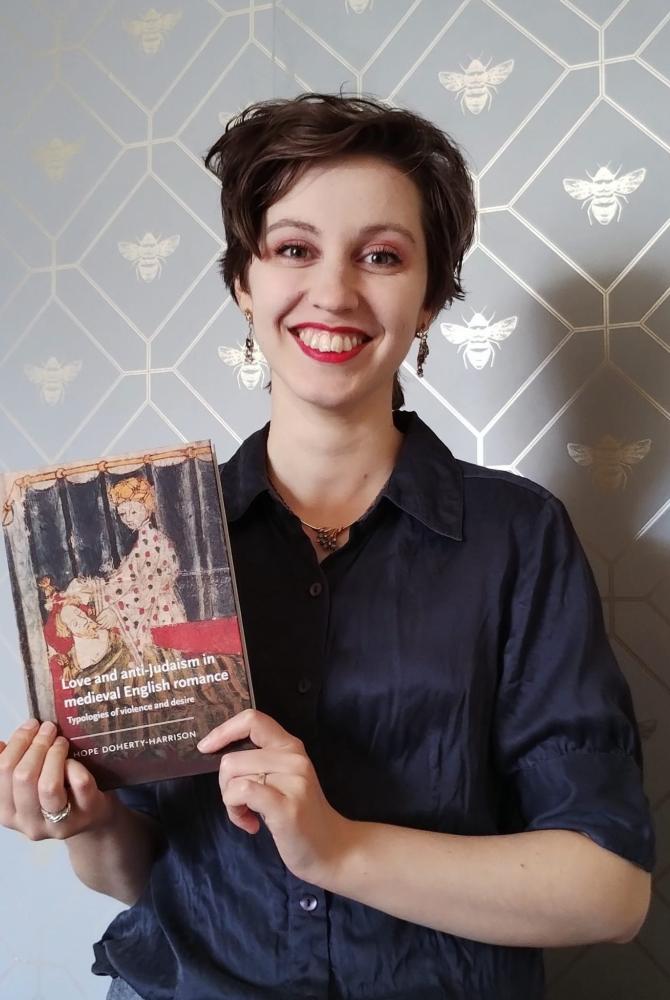 A person with short dark brown hair, smiles tat the camera. They are holding a book in front of them, which they have written.