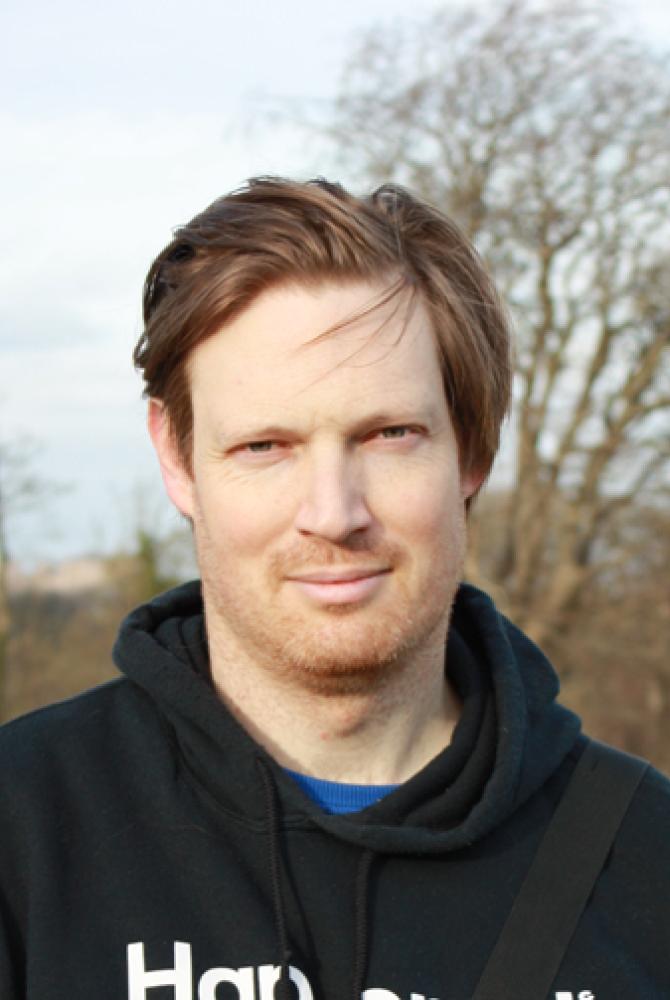 Dr Tom Mudd stands outside in a black hooded jumper. There are trees in the background.