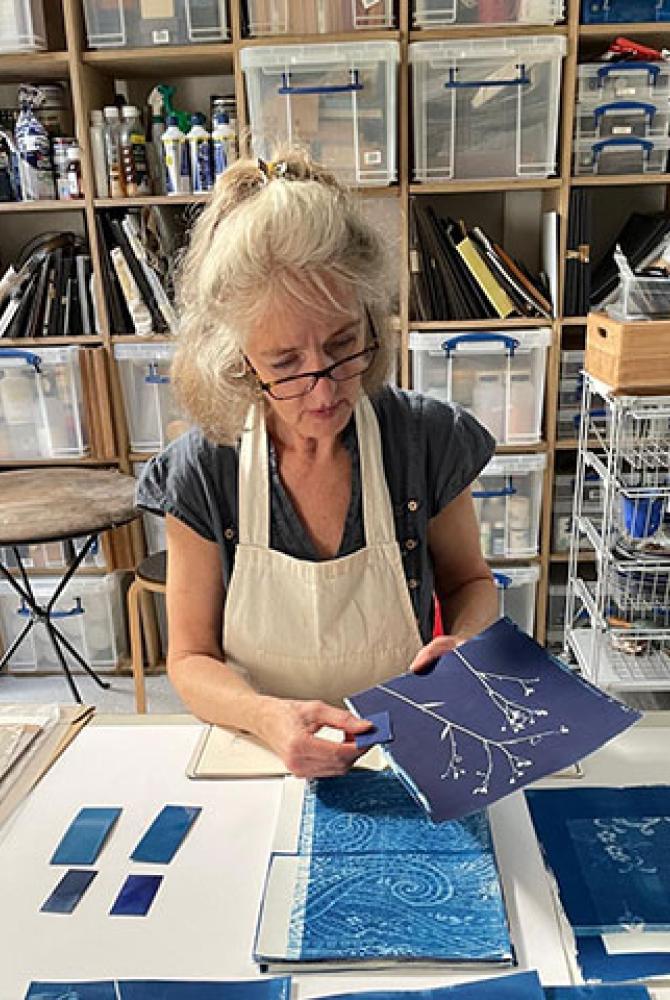 Susan Cross sitting at a table in a studio, with cyanotype prints of plants.