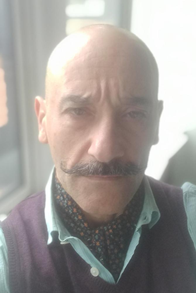 A person with a shaved head, a moustache twirled up at the ends, looking straight to the camera.