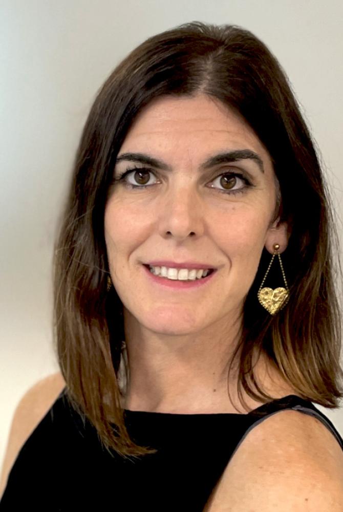 Pilar smiles to the camera. Their hair is dark brown and shoulder length and they wear a black strappy top and gold heard earrings.