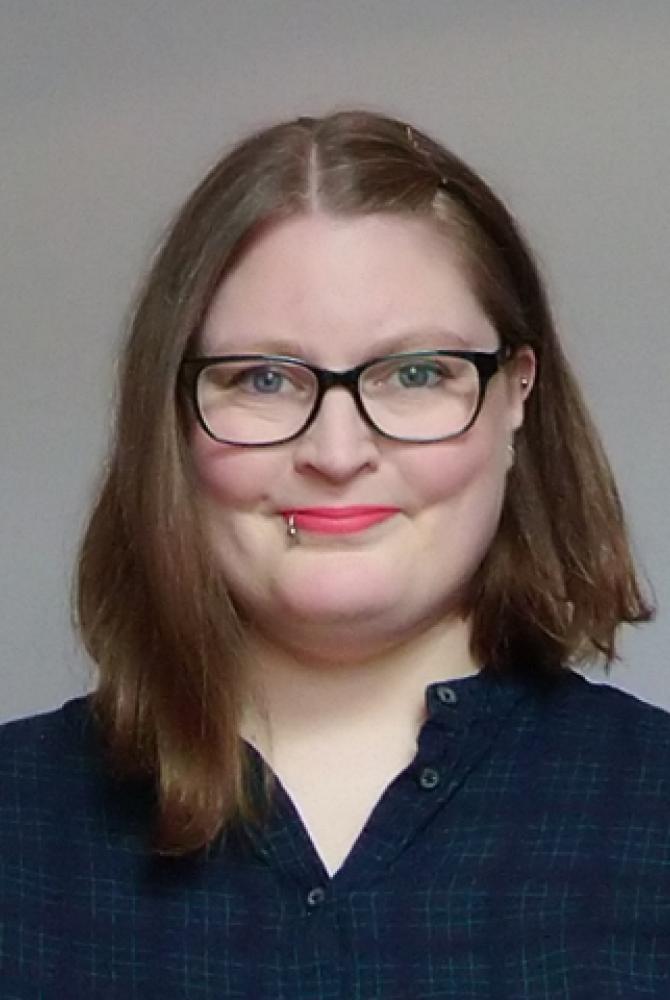 Suzanne black looks straight to camera. They have short, mid brown hair and wear black glasses and a black shirt.