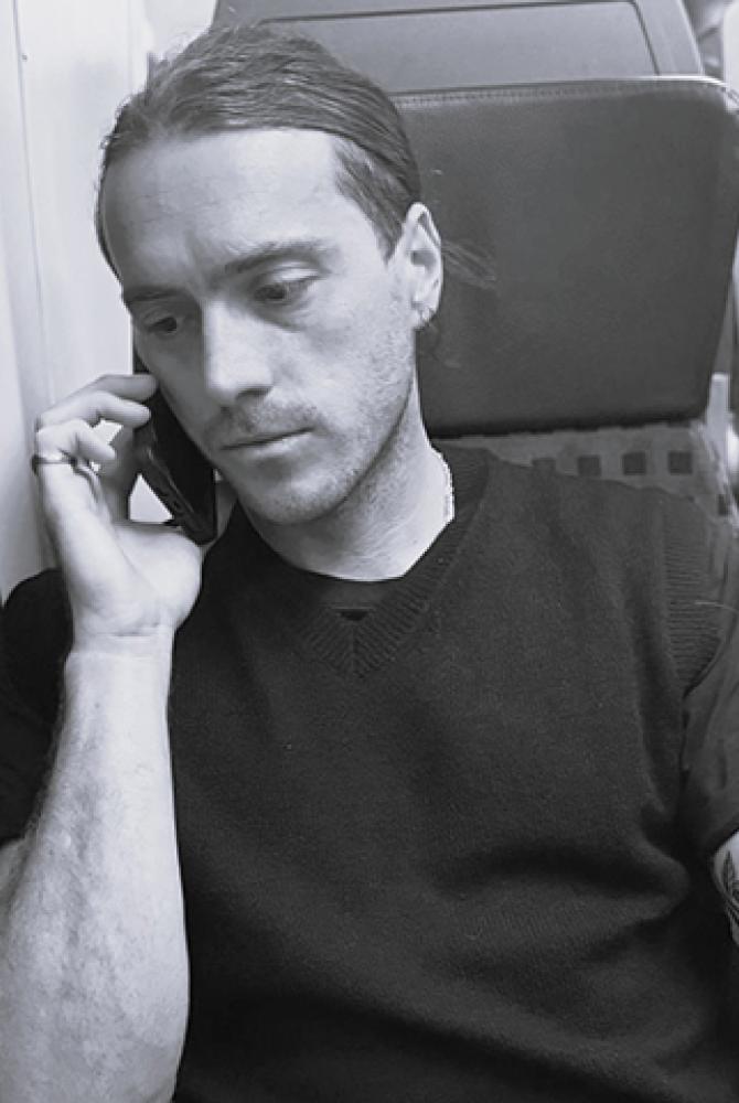 A black and white photo of Ian sitting on a train, holding a phone to his ear.