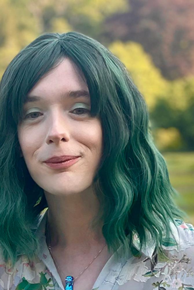 Dr Merlyn Seller has green hair and is wearing a floral blouse. They are standing in a park, smiling to the camera.