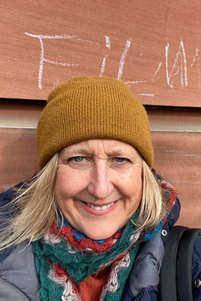 Tracey stands outside the Main Building ECA, under a chalk sign that reads 'Film'. They wear a waterproof coat, scarf and mustard beanie hat.