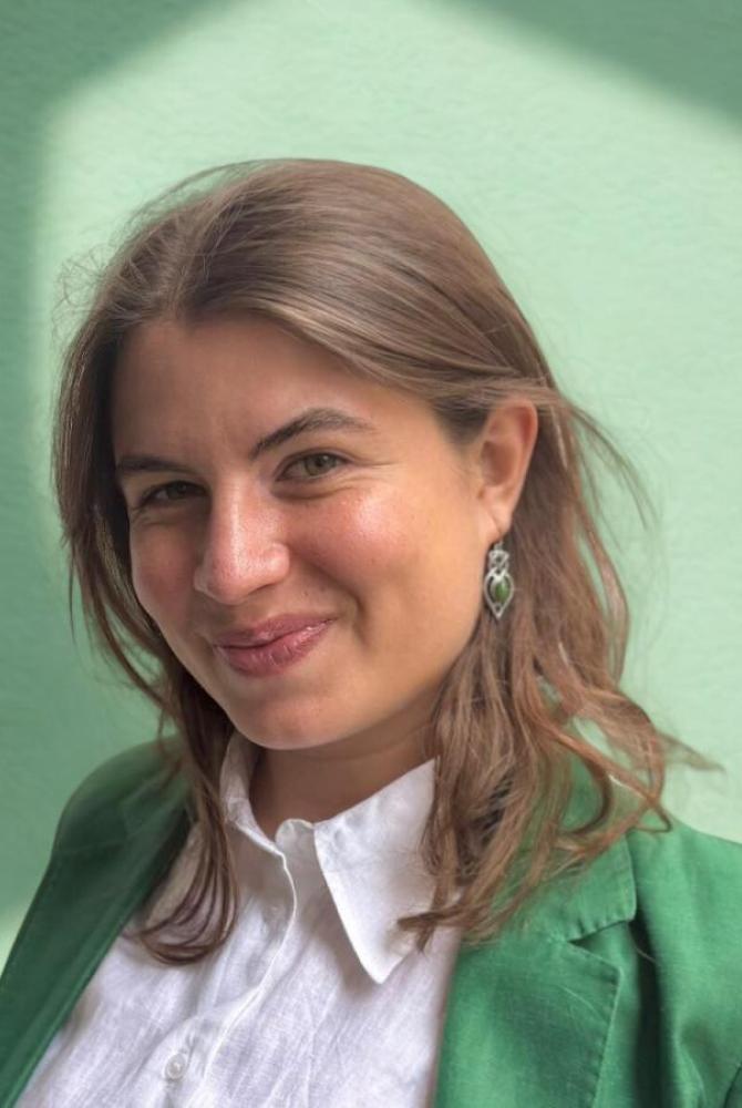 Prune smiles to the camera. She wears a white shirt and a green jacket.
