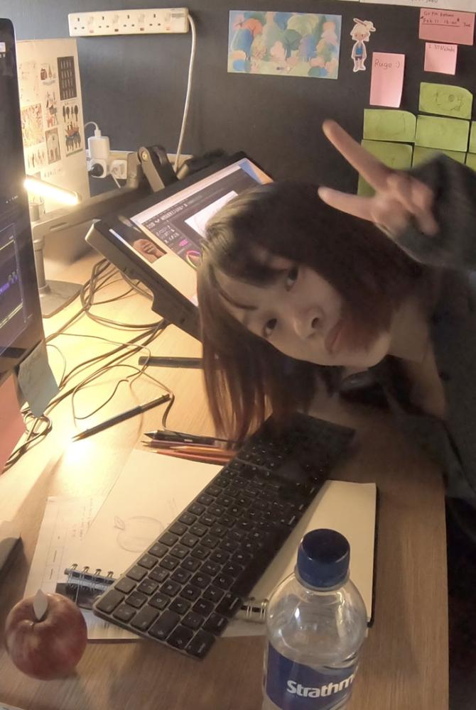 Ruge sits at her desk in front of a large monitor. Her animation work is around her and she does the peace sign to camera.