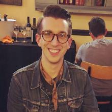 A person with short brown hair, wearing glasses and a denim jacket, and smiling at the camera.
